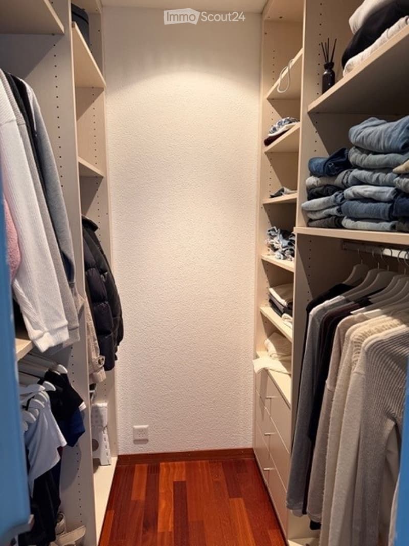 Well-organized walk-in closet with wooden flooring, built-in wardrobe, shelves, and neatly arranged clothing items.