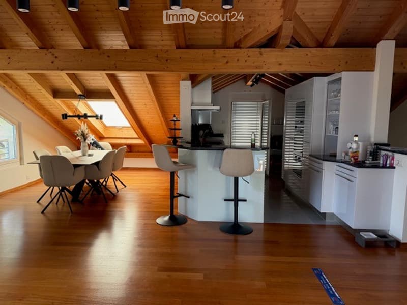 Bright, open-plan living and dining area with wooden floors and exposed wooden beams. Features a dining table with chairs, a kitchen island with bar stools, modern white kitchen cabinets, and a coffee machine on the counter.