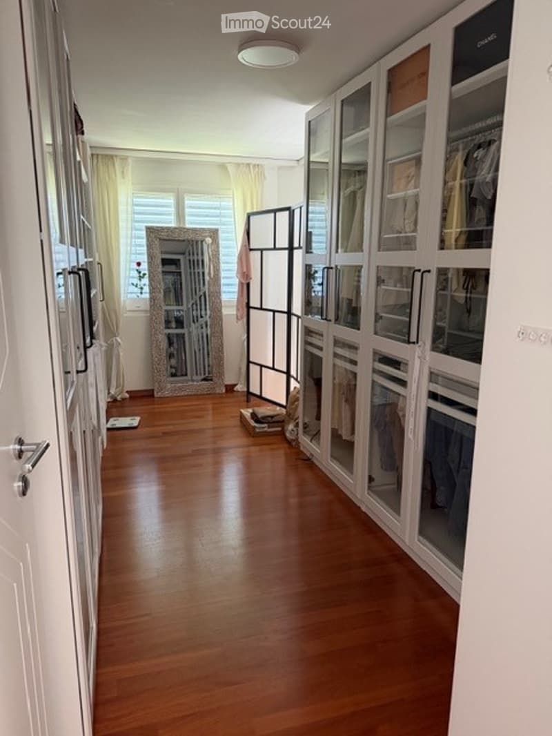 Spacious hallway with wooden flooring, large mirrored closet, and built-in wardrobe with glass doors. Natural light from windows and a ceiling light illuminate the space.