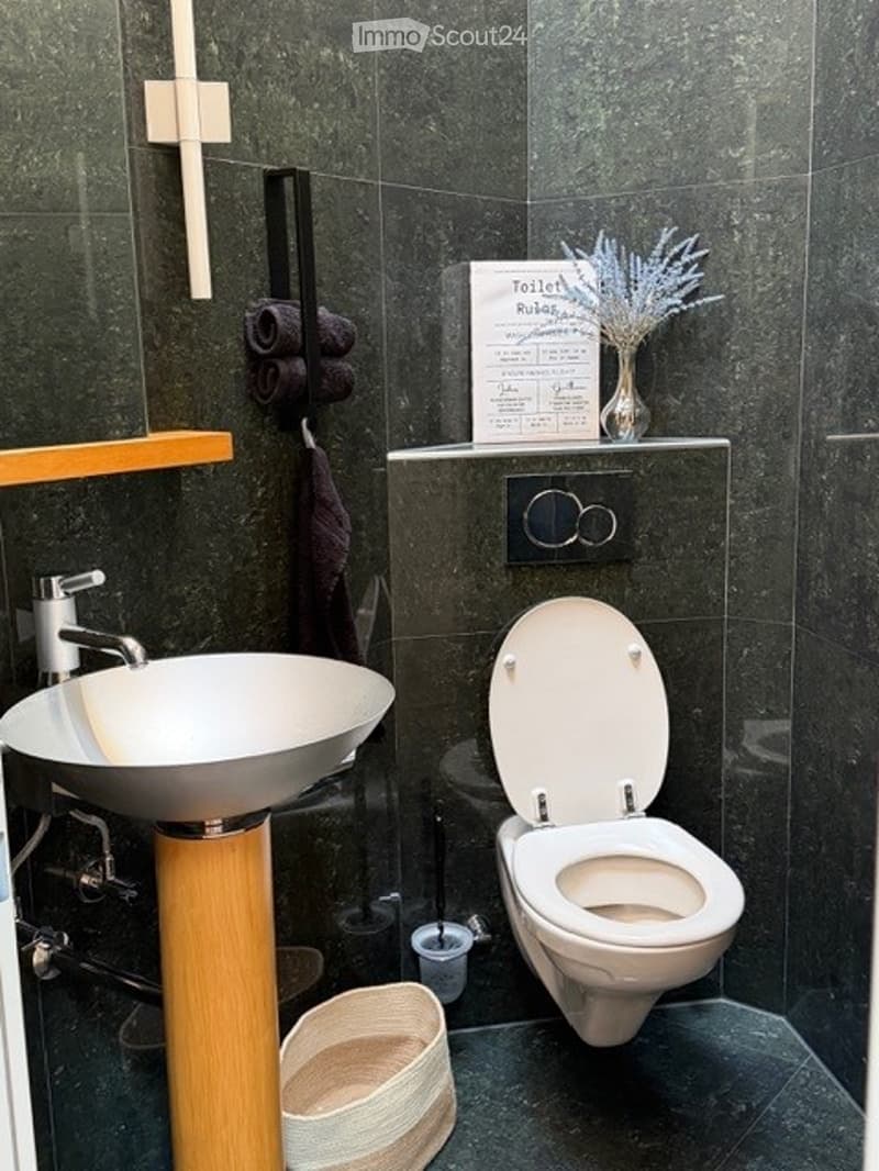 Modern bathroom with dark green tiles, a white sink with a wooden stand, a toilet, a towel rack with towels, a small shelf with a vase of flowers, and a decorative sign.