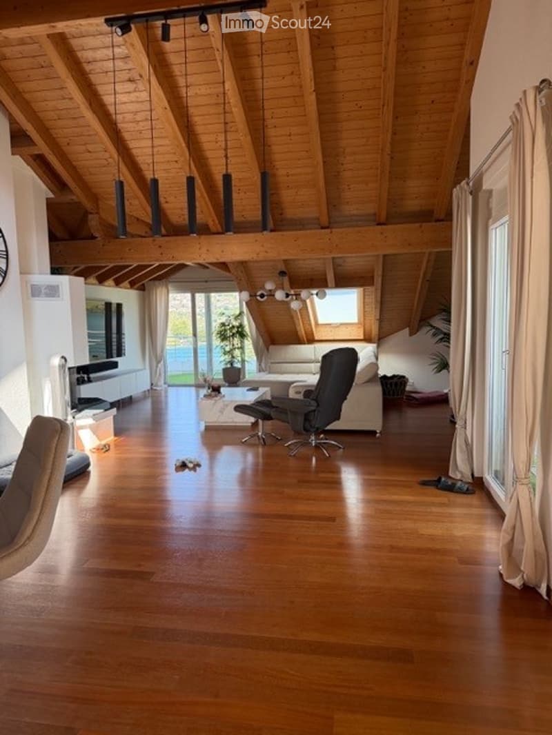 Spacious living room with exposed wooden beams, hardwood flooring, large windows, and a view of the garden. Features a white sofa, coffee table, chairs, and a television.