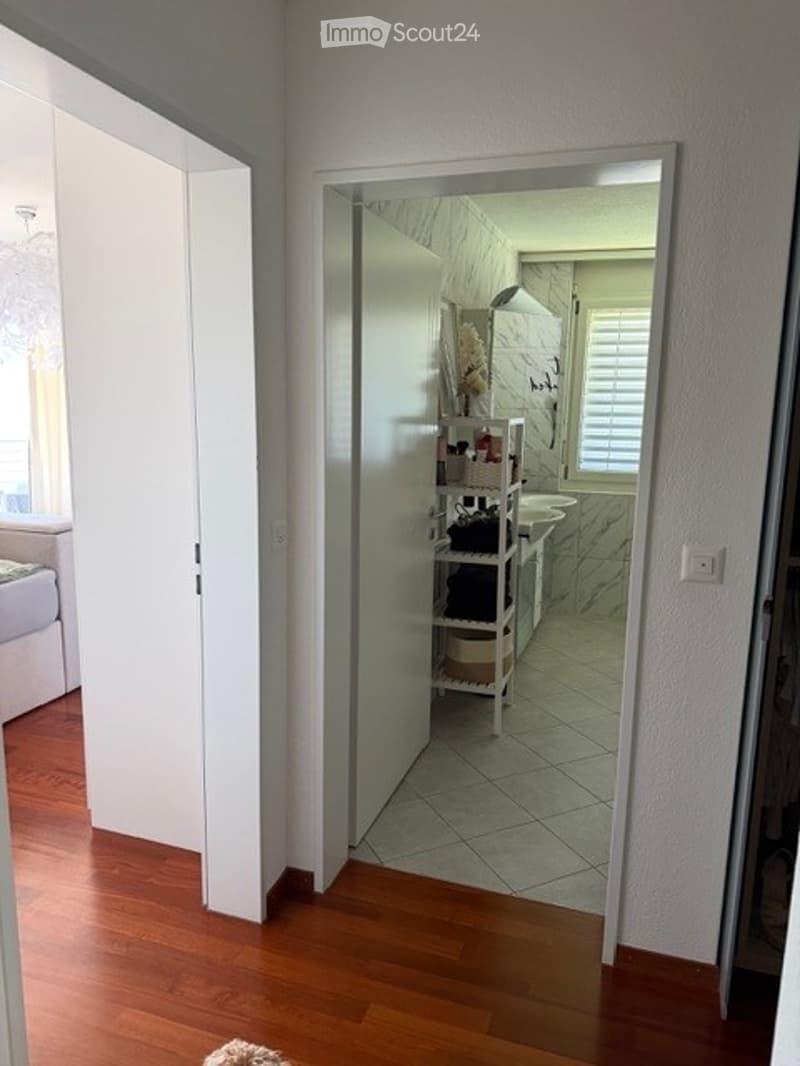 A hallway with wooden flooring leading to an open bathroom area. The bathroom features marble tiles, a sink, and a small shelf with various items.