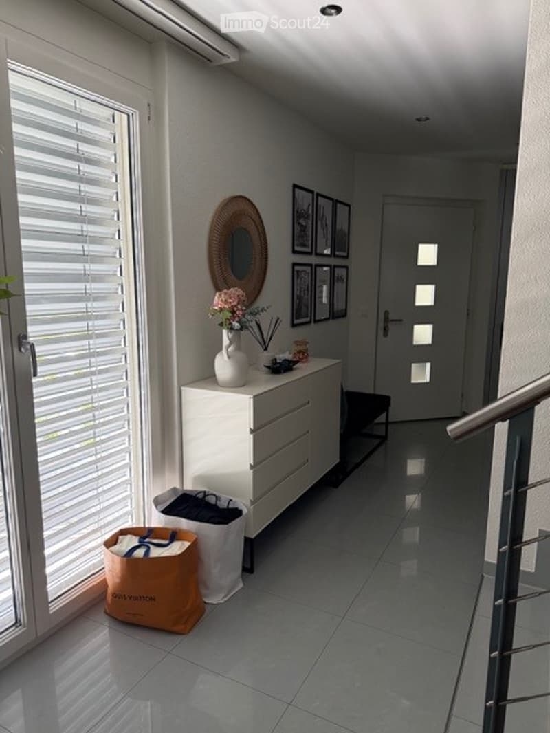 Modern hallway with white walls, grey tiled floor, white cabinet with drawers, decorative mirror, framed artwork, and a vase with flowers. An orange bag is placed on the floor near the window with blinds.