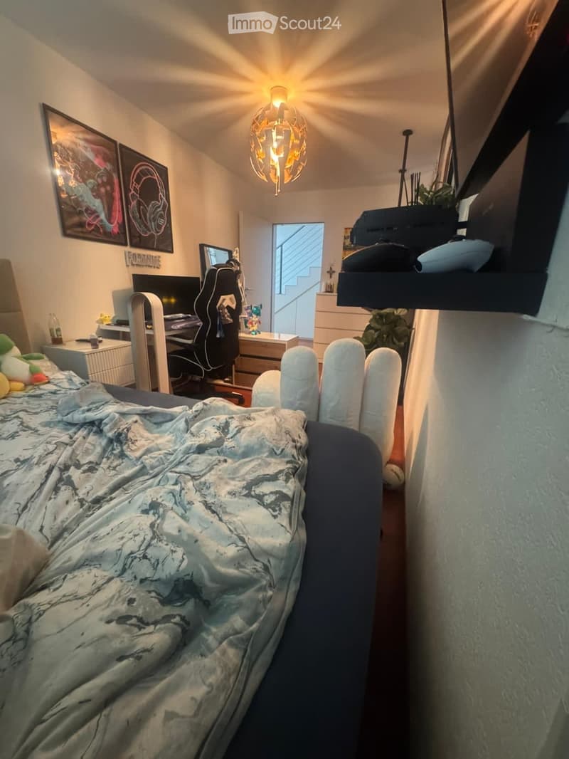 A cozy bedroom with a large bed, a desk with a computer, a guitar, and a small plant. The room features a modern lamp and a view of a staircase through an open doorway.