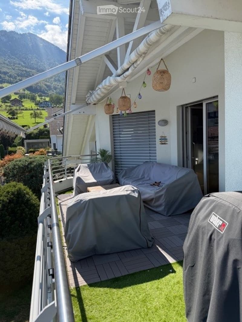 Balcony with covered seating area, covered furniture, hanging baskets, and scenic mountain view.