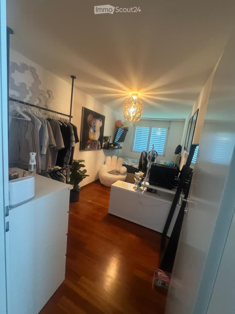 A furnished room with wooden flooring, a white chair, a small table, a screen, a plant, a lamp, and a picture on the wall.