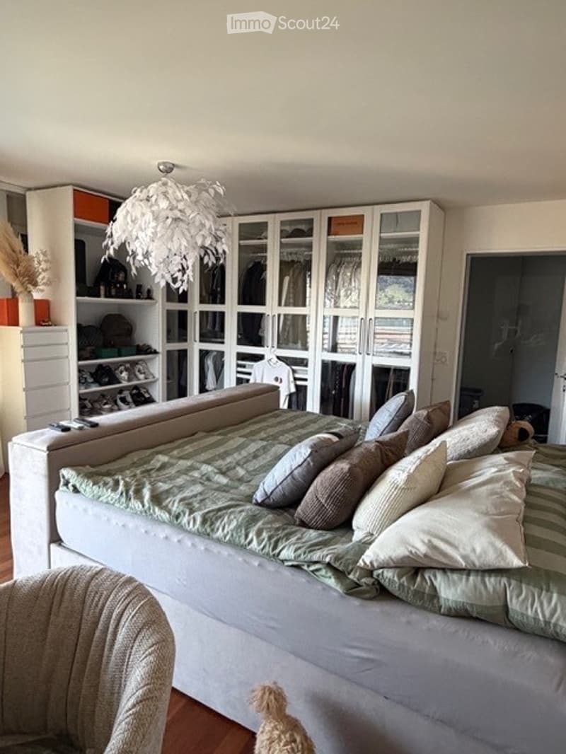 A modern bedroom with a large sofa bed covered in green and white striped bedding, multiple pillows, a wardrobe with glass doors, shelves filled with items, a decorative hanging lamp, and a small chair in the corner.