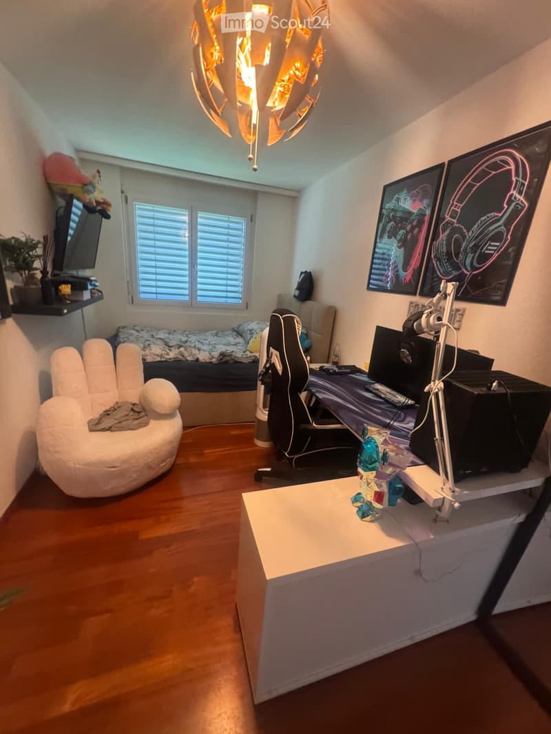 A modern, multi-functional room with a cozy sofa, a gaming chair, a desk setup with a laptop and multiple screens, and decorative elements like picture frames and a plant. The room features wooden flooring and large windows with blinds.