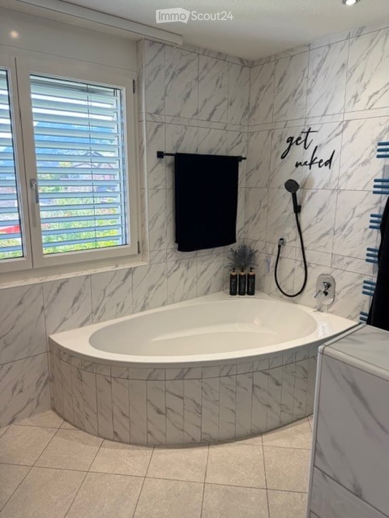 Modern bathroom with marble tiles, a large corner bathtub, a black towel, and a window with blinds.