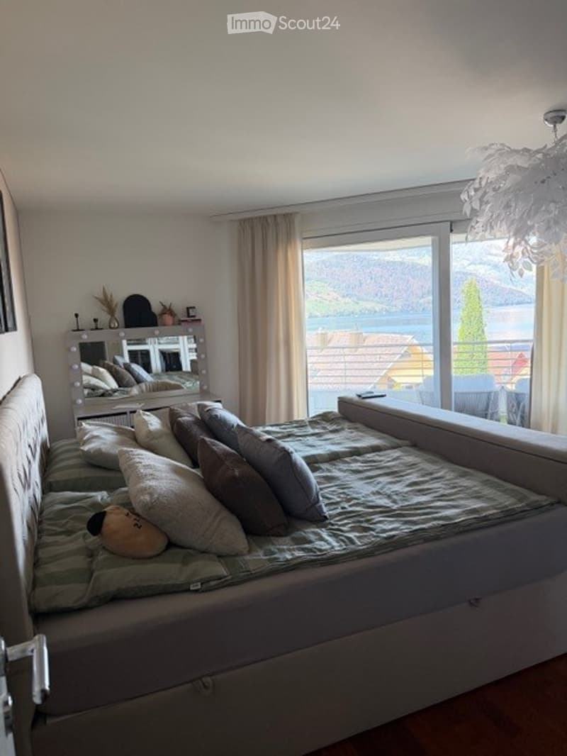 Modern bedroom with a large bed, multiple pillows, a vanity mirror, and a scenic view of a lake through large windows.