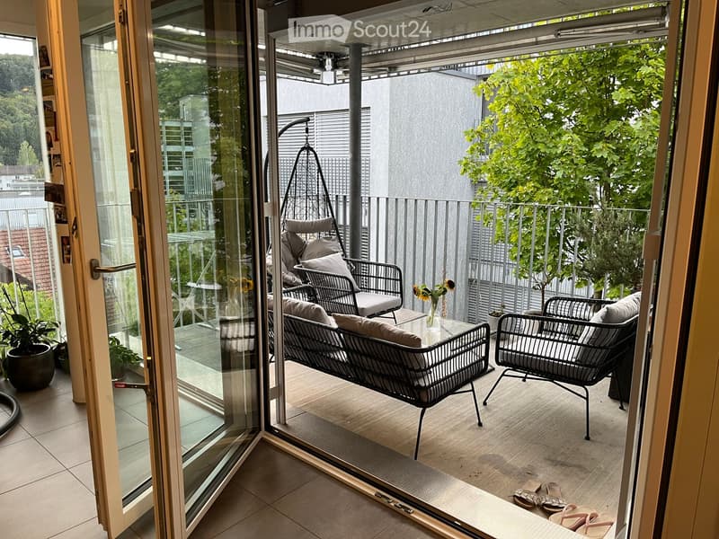 Balcony with a sofa, chairs, a small table, a vase with flowers, and potted plants. The balcony has a wooden floor and is enclosed with a metal railing.