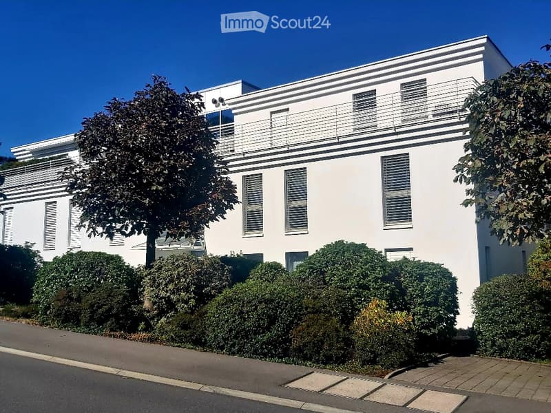 Modern two-story white building with large windows, balconies, and trimmed hedges in front.