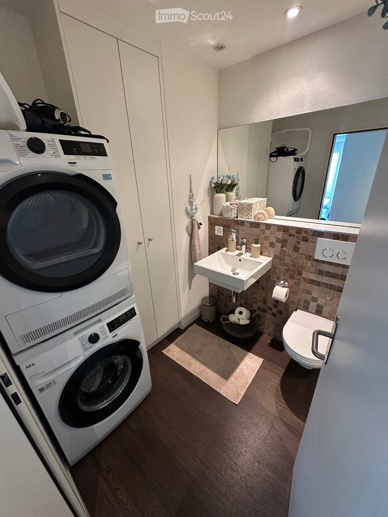 Modern bathroom with a sink, toilet, washing machine, dryer, and a mirror. The room features a tiled backsplash, a small window, and a wooden floor.