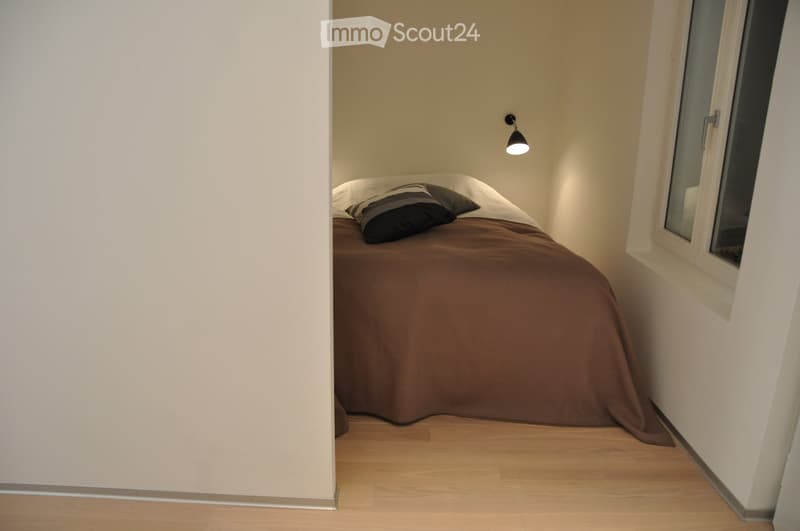 A small, cozy bedroom with a single bed covered in a brown duvet and pillows, positioned against a light-colored wall. The room features wooden flooring and a window with white frames on the right side.