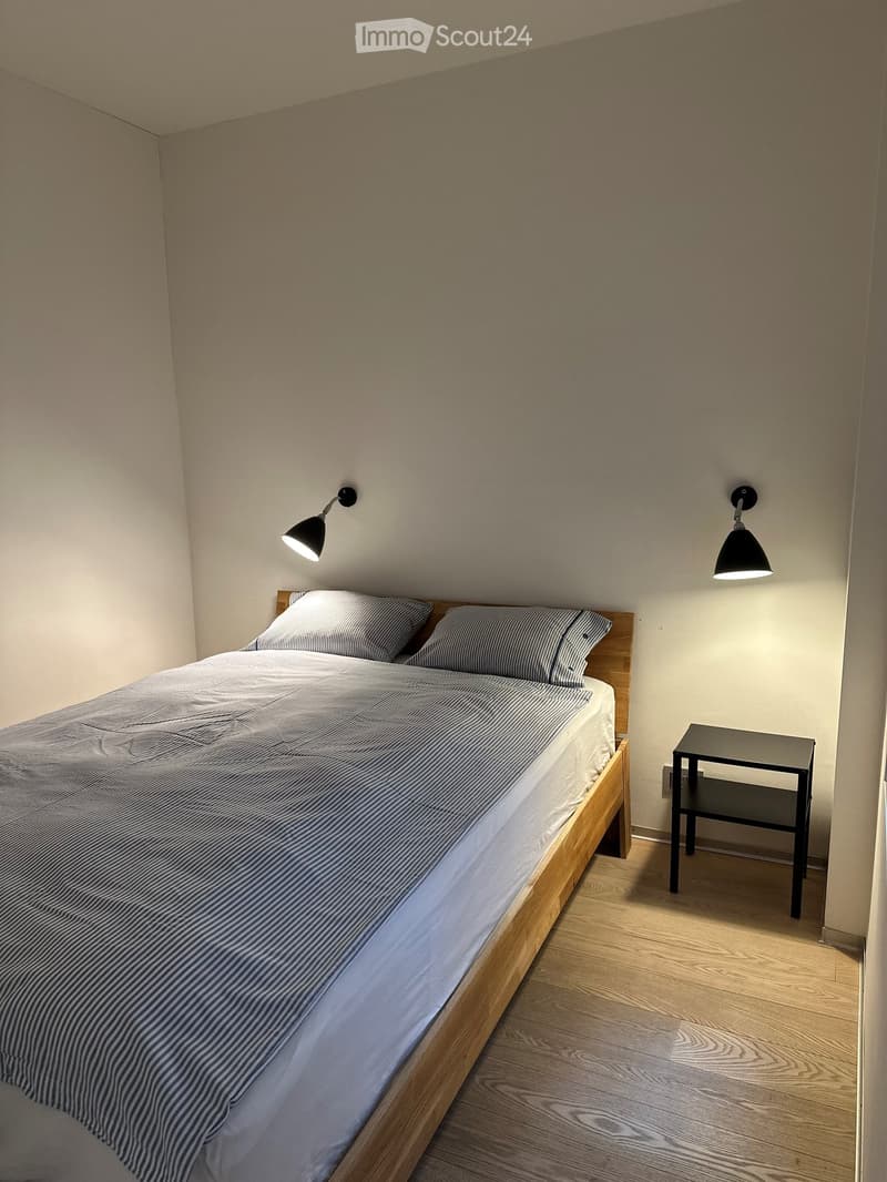 A small, modern bedroom with a wooden bed, striped bedding, a nightstand, and wall-mounted lamps.
