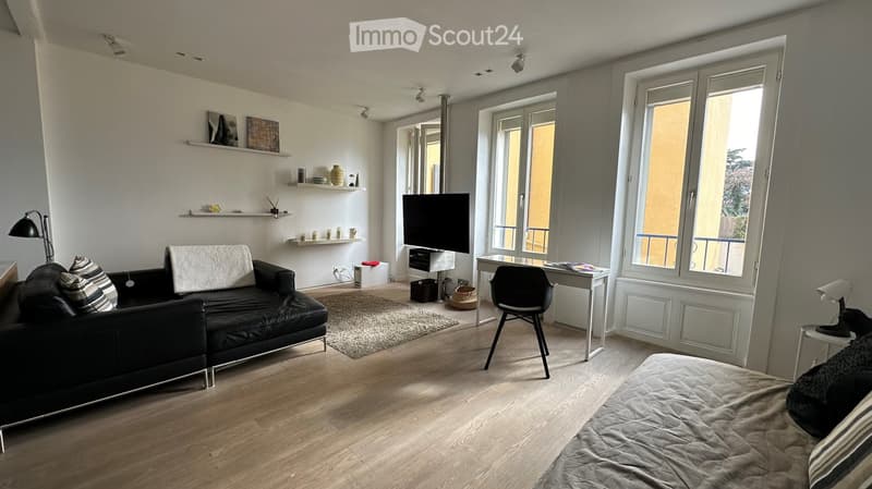 Modern living and sleeping area with a black leather sofa, a small desk with a chair, a television, and a bed with a grey duvet. The room features wooden flooring, large windows, and decorative shelves with vases and candles.