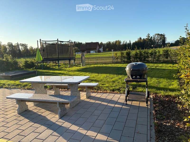 Outdoor patio with stone table, benches, grill, and umbrella, surrounded by a grassy lawn and trees.
