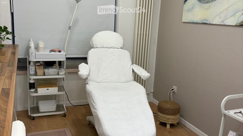 A well-lit, clean treatment room with a white treatment chair, a mobile cart with medical supplies, a small wicker stool, a painting on the wall, and a small plant on a wooden shelf.
