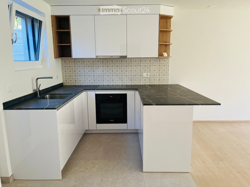 Modern kitchen with white cabinets, black countertops, a sink, and an oven. Features a window with a view and a wooden shelf on the right side.