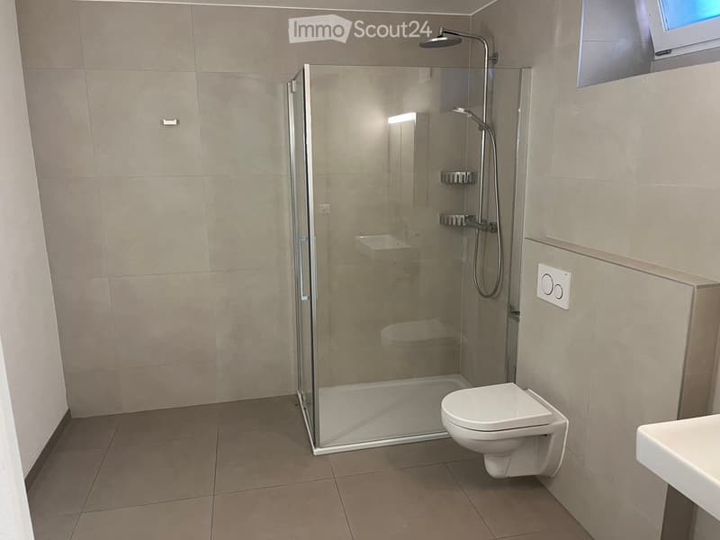 Modern bathroom with a glass-enclosed shower, toilet, and sink. Light-colored tiles and a sleek design.