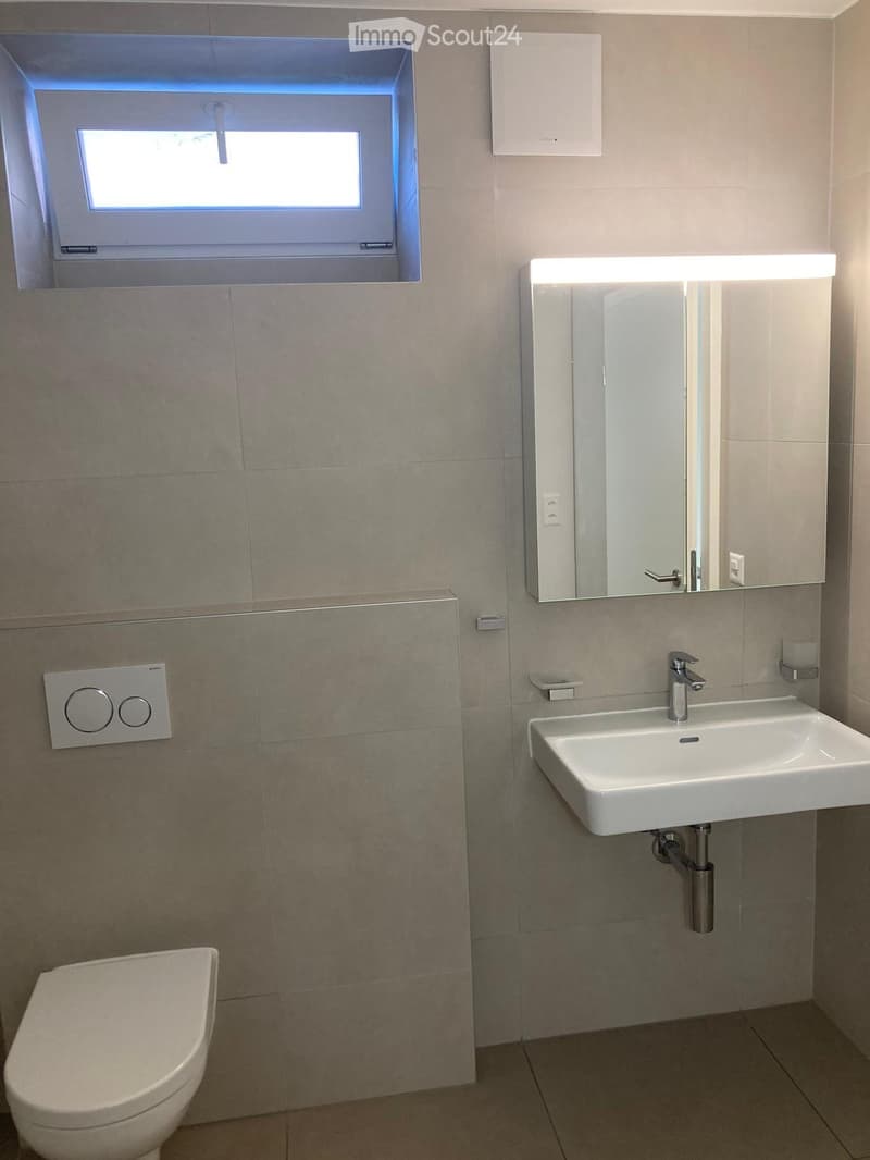 Modern bathroom with a white sink, mirror cabinet, and toilet. Features a window above the sink and tiled walls.