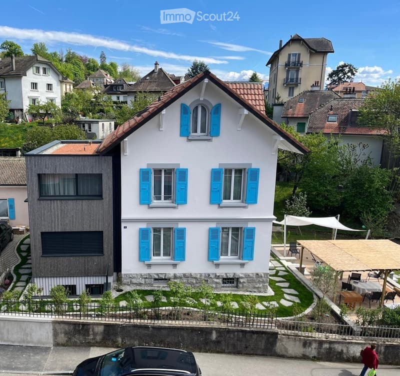 Two-story house with white walls, blue shutters, and a red-tiled roof. The house features a modern extension with dark wooden panels and large windows. The front yard includes a small garden with stepping stones and a covered outdoor seating area with tab