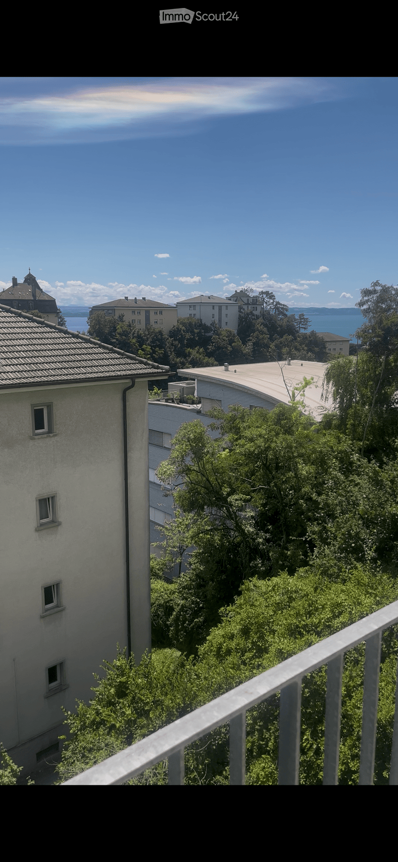 Balcony overlooking a lush green area with trees, featuring a metal railing and a view of neighboring buildings and a body of water in the distance.