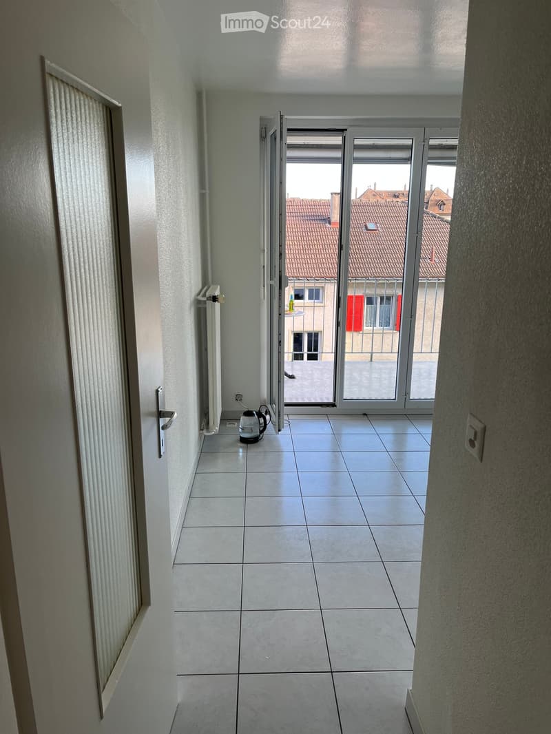 Small tiled room with a radiator, electric kettle, and an open door leading to a balcony with a view of rooftops.
