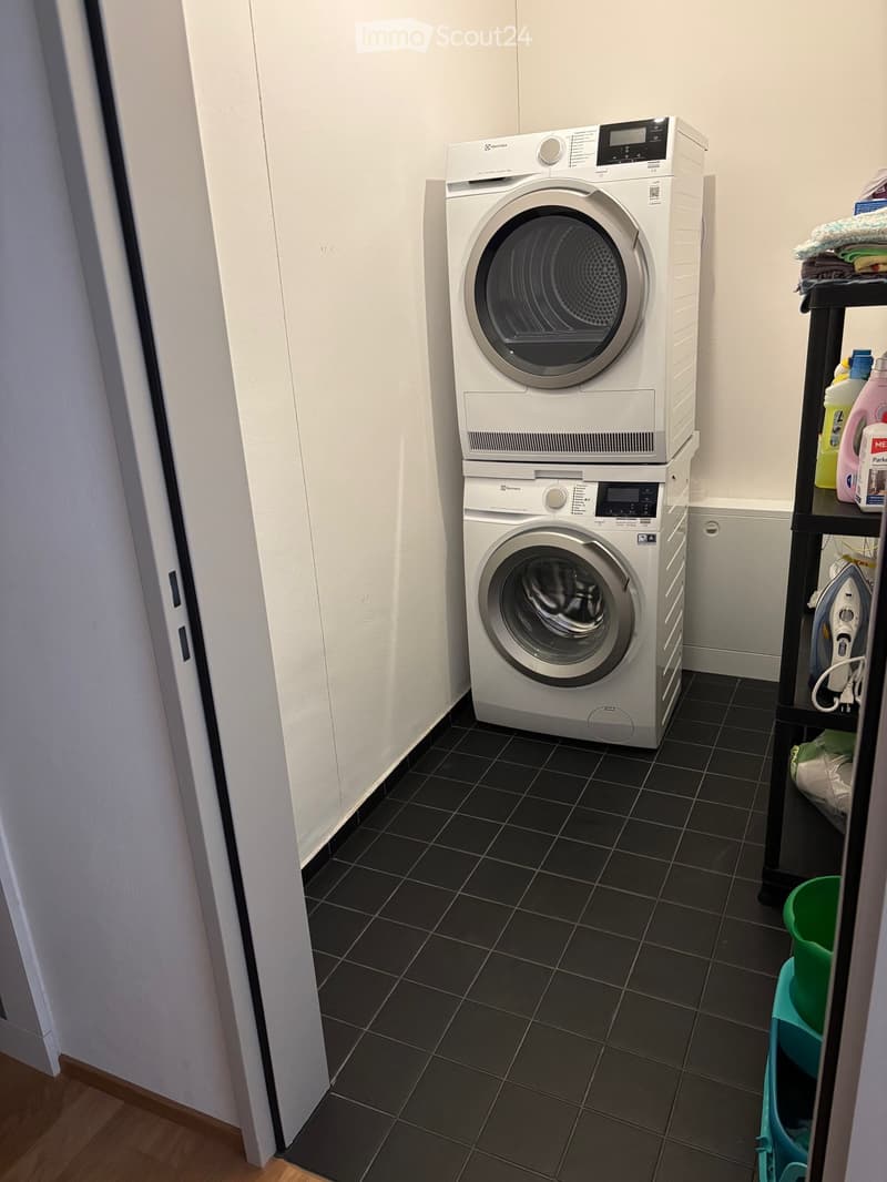 A small bathroom with a washing machine and dryer stacked vertically, a shelf with cleaning supplies, and a small ironing board.