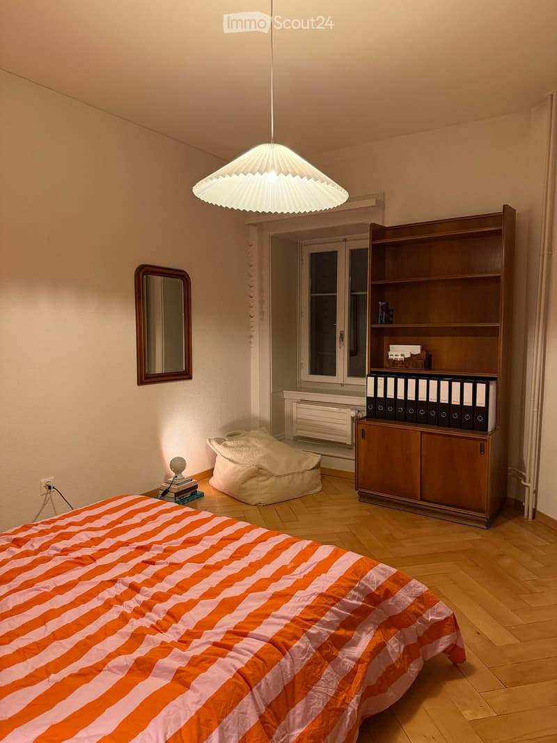 A well-lit bedroom with a wooden floor, a bed with an orange and white striped duvet, a bookshelf filled with binders, a round mirror on the wall, a small table with books and a lamp, and a hanging pendant light.