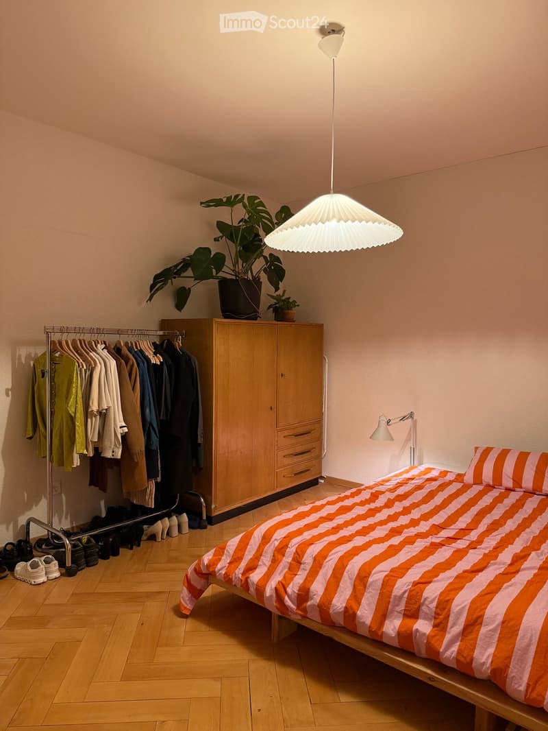 A modern bedroom with a wooden bed, orange and white striped bedding, a large wardrobe, a clothes rack, and a hanging lamp.