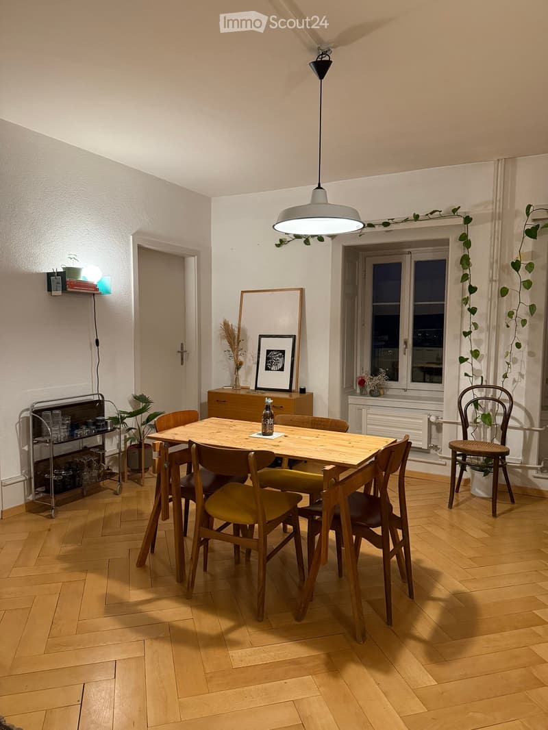 Bright kitchen with wooden flooring, a central wooden dining table surrounded by chairs, pendant light fixture, cabinet, picture frame, shelf with glassware, and potted plants.