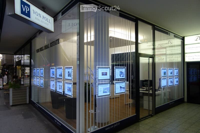 Exterior view of a real estate office with glass windows displaying property listings, signage for "Von Poll Real Estate", and interior furniture visible through the glass.