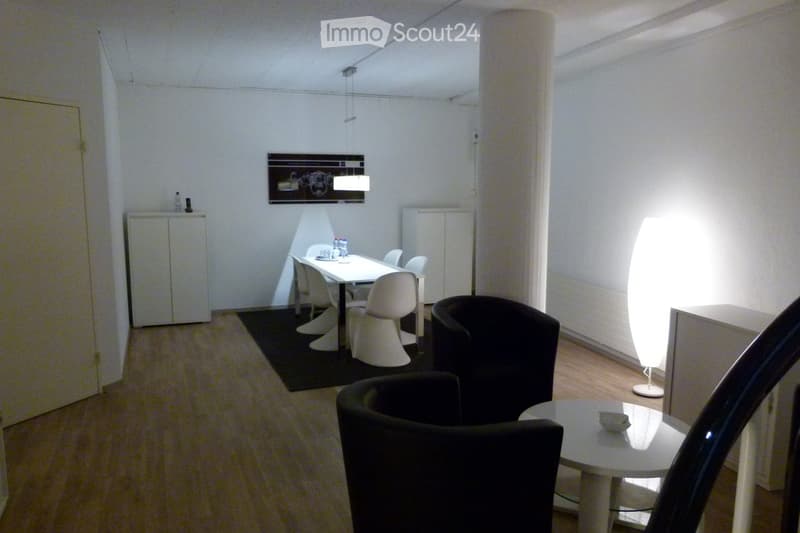 Modern, minimalist room with a white dining table and chairs, a black armchair, a tall floor lamp, and a small cabinet with bottles on top.