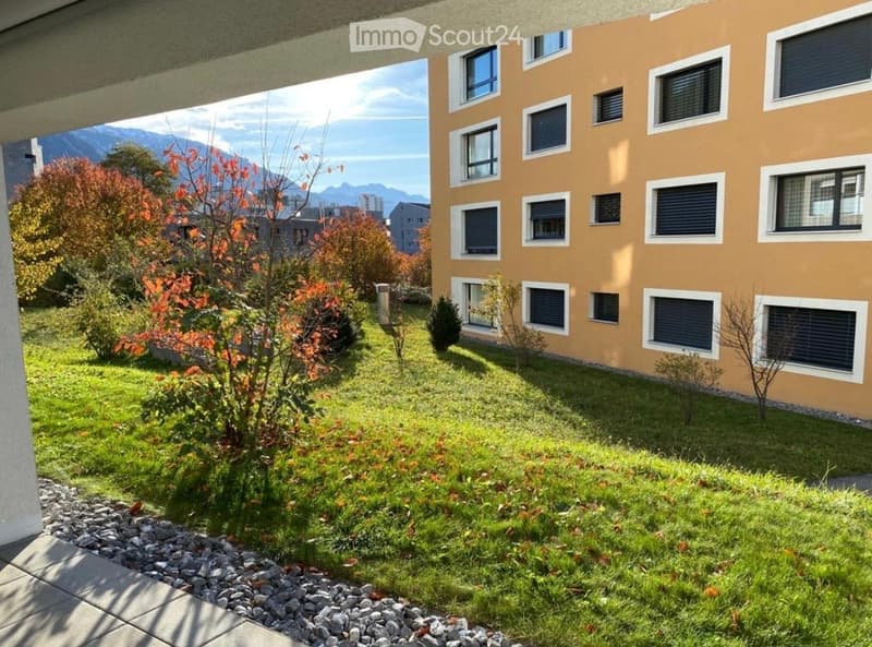 Well-maintained garden area with green grass, small shrubs, and a view of mountains in the background. The building has a light brown facade with multiple windows.