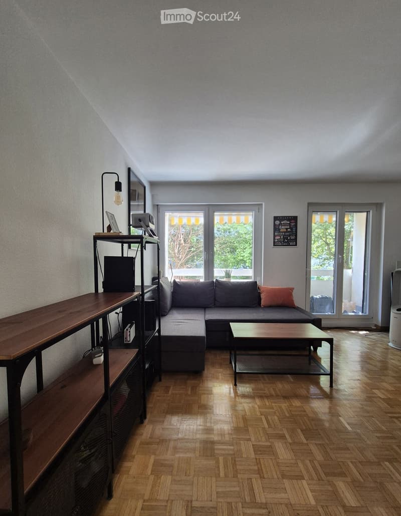 Living room with wooden flooring, grey sectional sofa, coffee table, shelf with electronic devices, and a lamp.