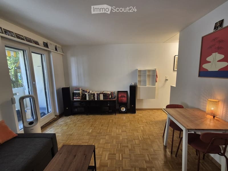 Living room with wooden herringbone flooring, large window with balcony access, sofa, coffee table, dining table with chairs, bookshelf, lamp, and speakers.