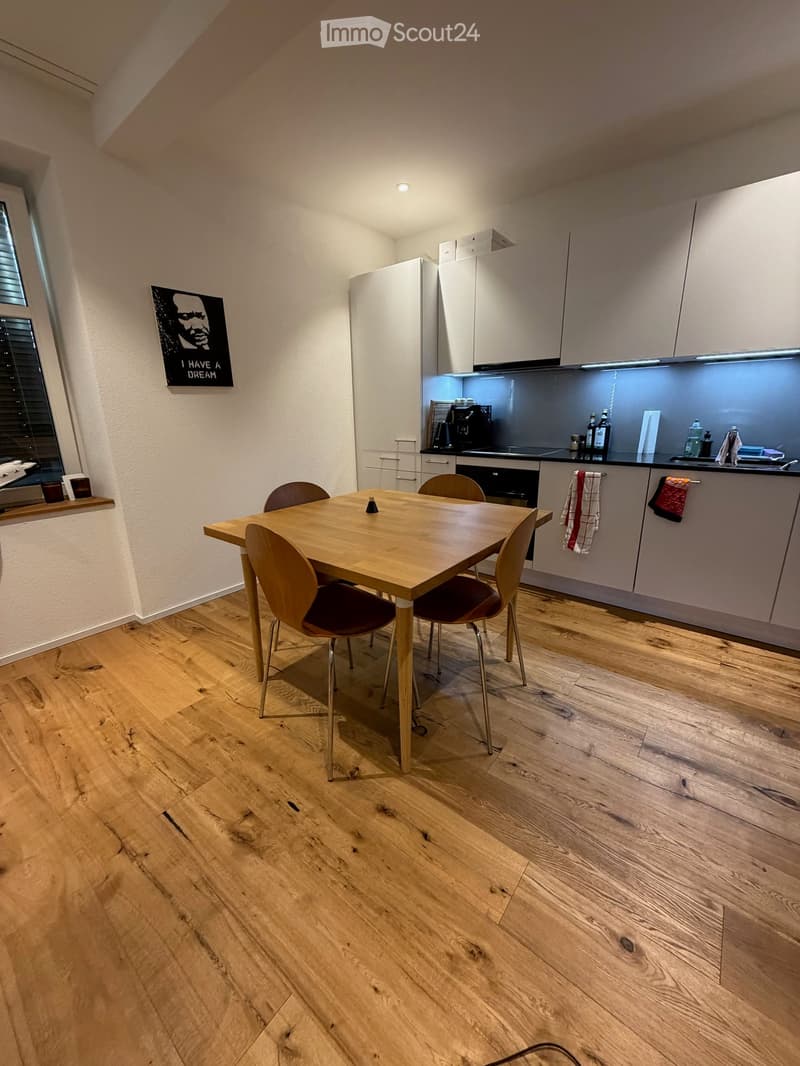 Modern kitchen with light wood flooring, white cabinets, a dining table with four chairs, and various appliances including a refrigerator, oven, and coffee machine.