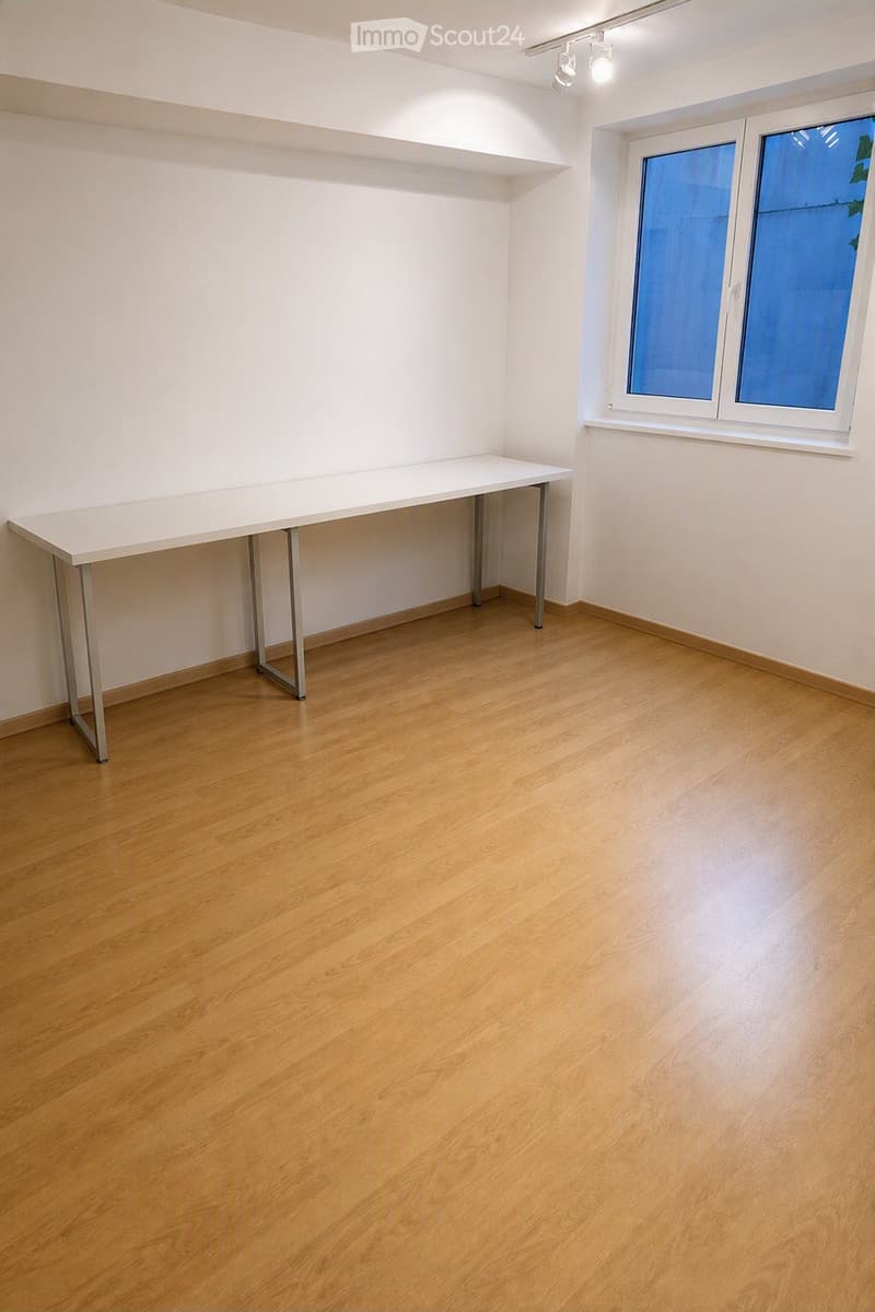 Empty room with light wooden flooring, white walls, and a single white table against one wall. A window with blue-tinted glass is visible on the right side.
