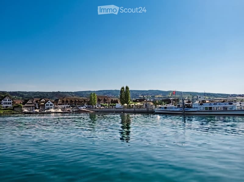 A serene lakeside view with calm water, a docked boat, and traditional houses along the shore under a clear blue sky.