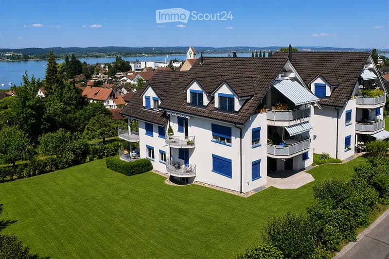 A modern two-story house with white walls, blue shutters, and multiple balconies overlooking a scenic lake and town.