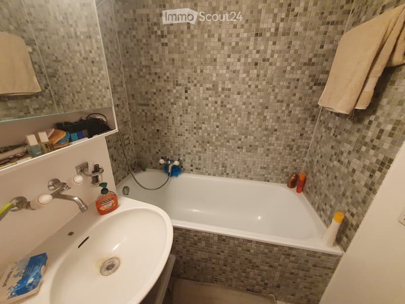 Bathroom with a white sink, bathtub, and mosaic tile walls. A mirror above the sink, and towels hanging on the side.