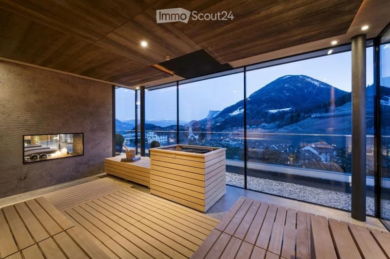 Modern sauna room with wooden benches, a wooden sauna, a fireplace, and a potted plant, offering a scenic mountain view through large windows.