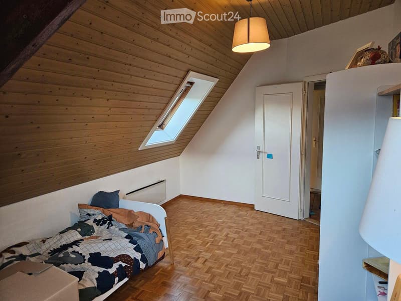 Cozy attic bedroom with wooden walls and herringbone flooring, featuring a single bed with colorful bedding, a pendant light, and a built-in shelf unit.