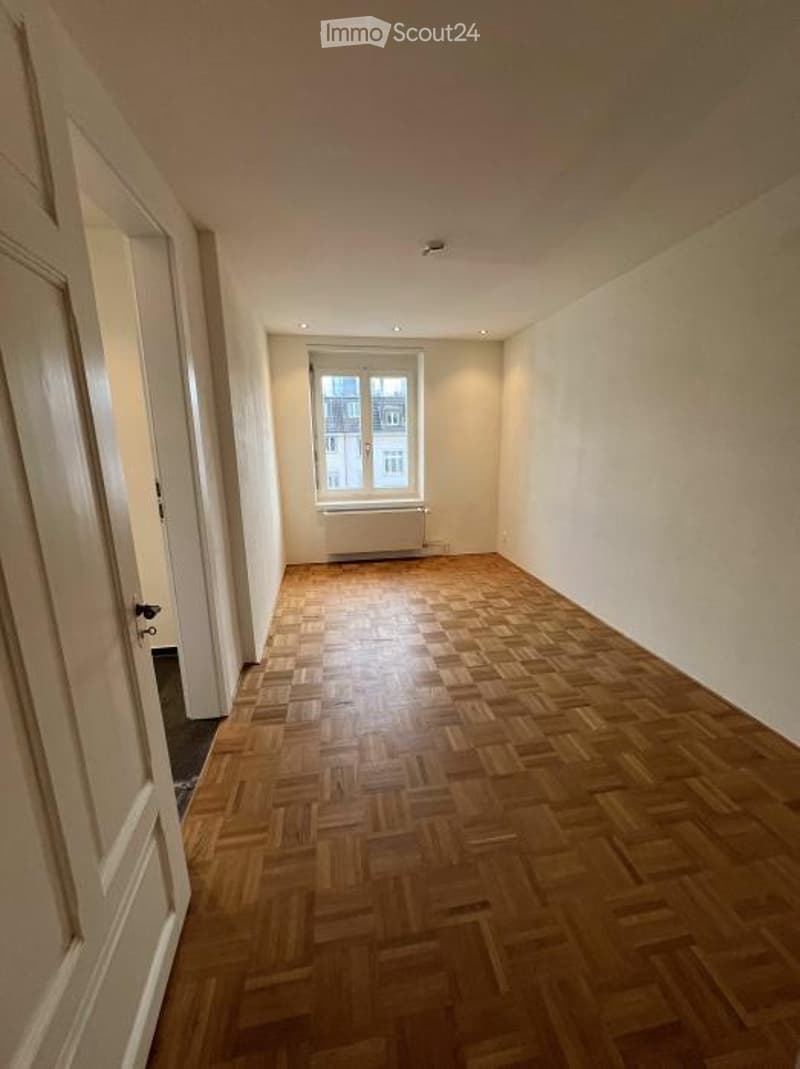 Empty room with wooden parquet flooring, white walls, and a single window with a radiator below it. The room has a door on the left side and recessed ceiling lights.