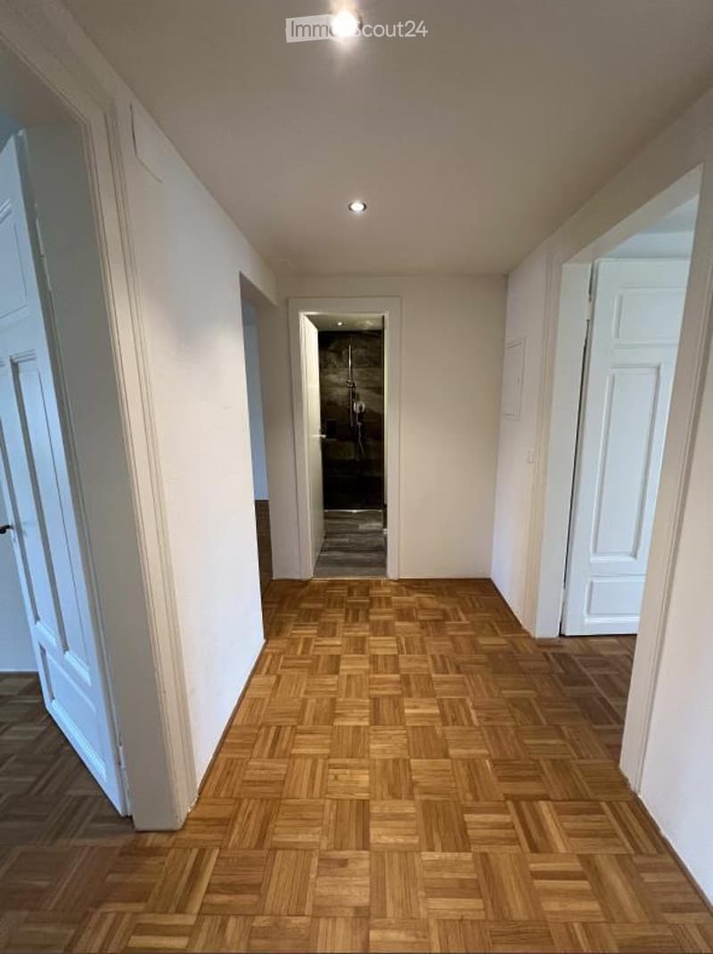 Empty hallway with wooden herringbone flooring, white walls, and recessed lighting. Doors lead to adjacent rooms, including a bathroom visible at the end.