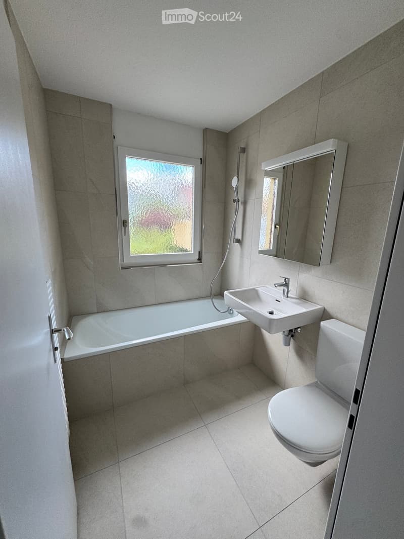 Modern bathroom with a bathtub, sink, toilet, mirror, and shower. Features a frosted window and tiled walls and floor.
