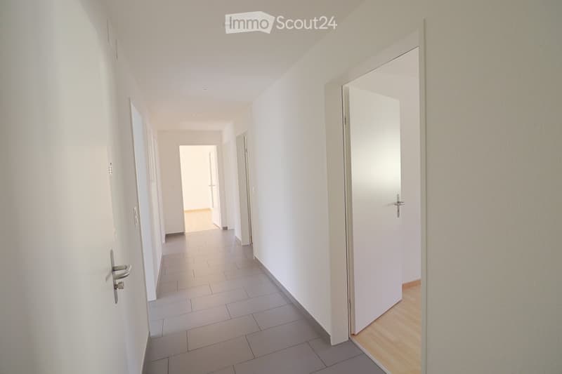 A clean, modern hallway with white walls, tiled floor, and several closed white doors. The hallway has recessed ceiling lighting and a wooden floor visible at the end of the corridor.