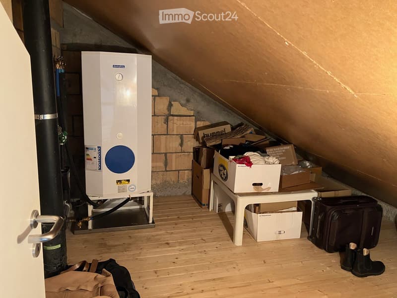 Attic space with wooden flooring, a white table holding boxes, a large white boiler, and personal items like a suitcase and backpack.