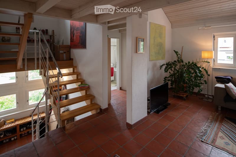 A modern, open-plan living area with a wooden staircase leading to an upper level. The space features a grey sofa, a flat-screen TV, a floor lamp, a large potted plant, and colorful artwork on the walls. The floor is tiled in terracotta, and there is a sm